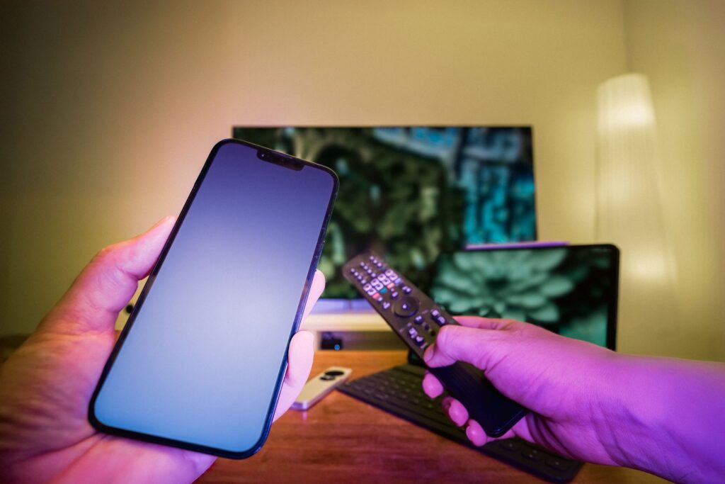 Dłonie ze smartfonem i pilotem przed telewizorem w nowoczesnym domu